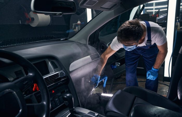 Worker of auto repair in mask bending and blowing chemical cleaning spray on car interior