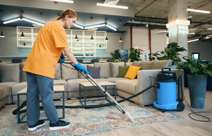 Cleaning company worker in a yellow uniform cleans the carpet with a professional vacuum cleaner in a rest area