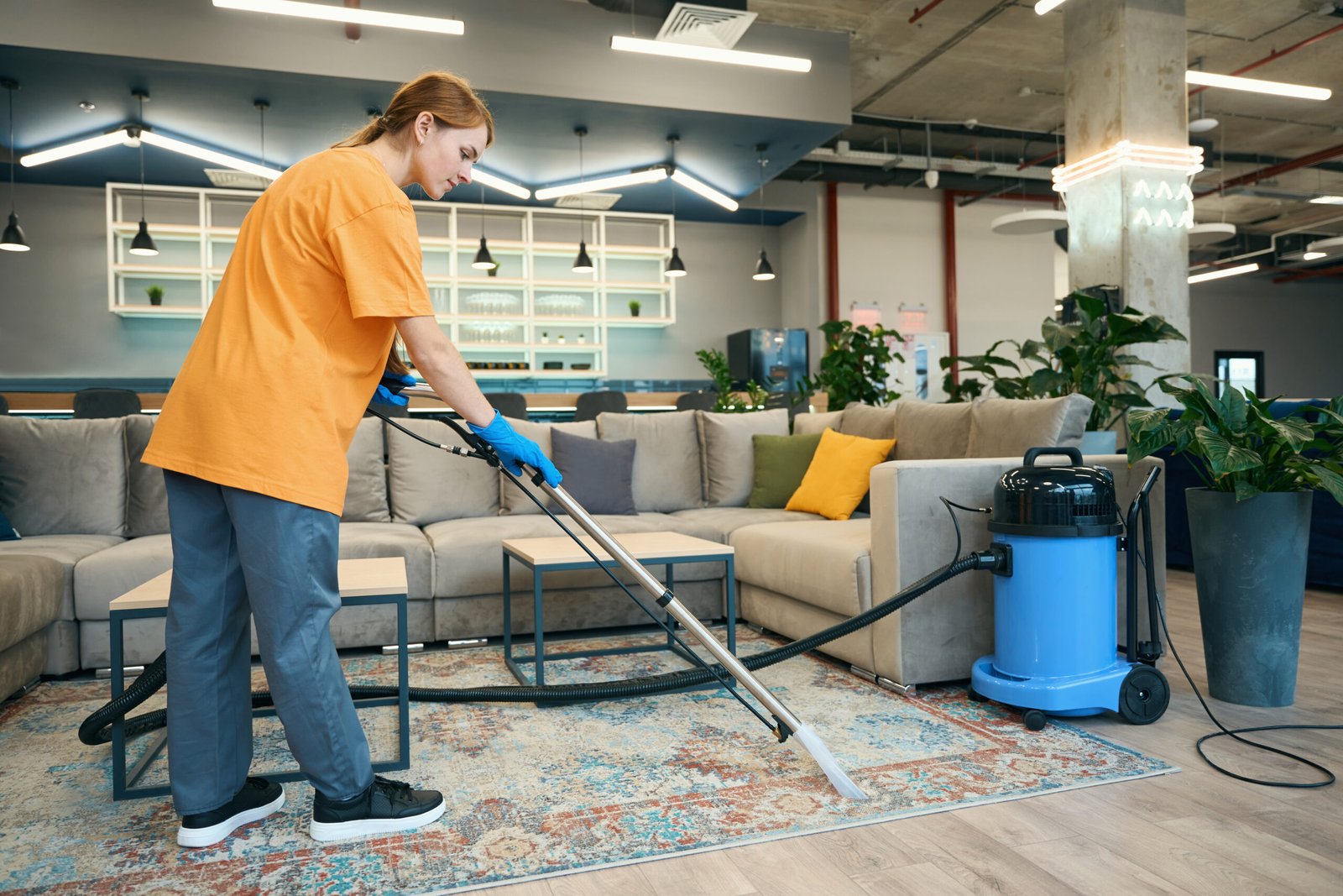 Cleaning company worker in a yellow uniform cleans the carpet with a professional vacuum cleaner in a rest area