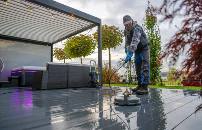 A person is cleaning a deck in a garden using a pressure washer. The sky is cloudy, and plants surround the area.