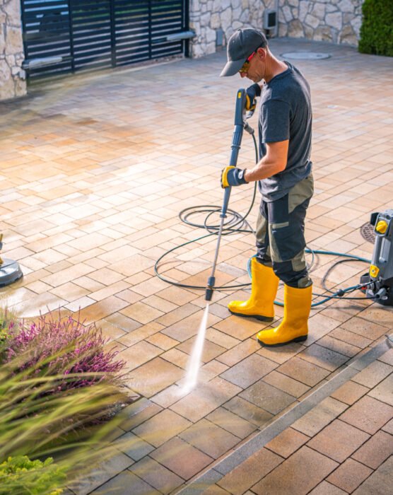 A man in yellow rubber boots operates a pressure washer to clean the patio stones in a garden. Lush greenery surrounds the area on a sunny day.