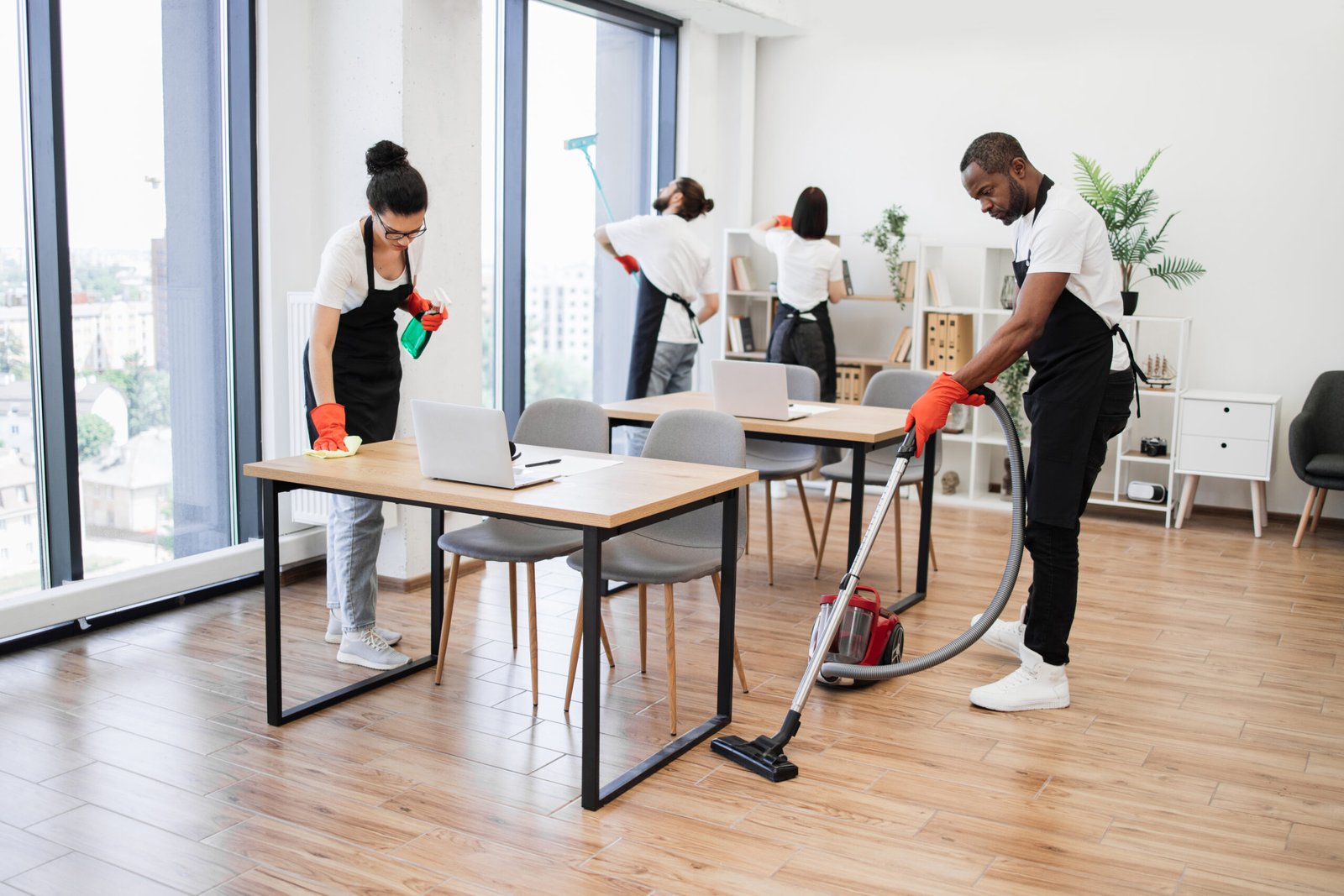 Team of young multicultural cleaners vacuums floor, wipes tables and gadgets, shelves, washes windows in spacious, bright, modern office. Professional cleaning company.