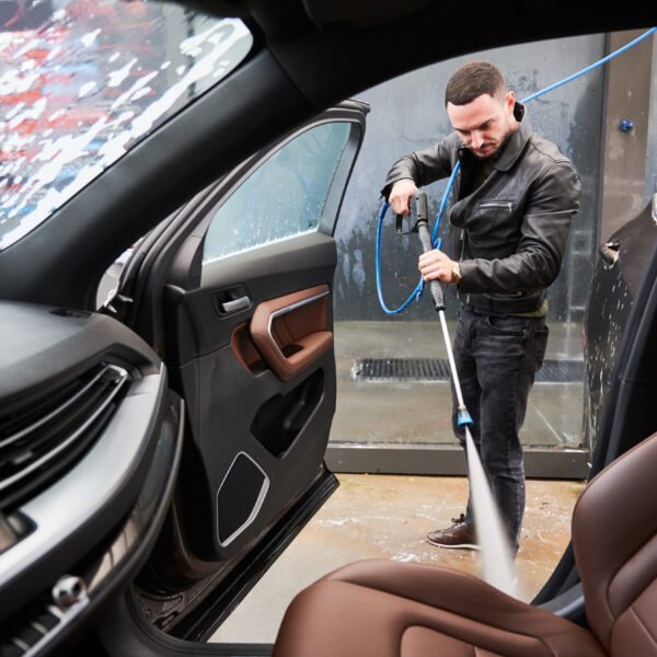 View from inside a car on young man washing car on carwash station outdoor. Handsome worker cleaning automobile, using high pressure water.