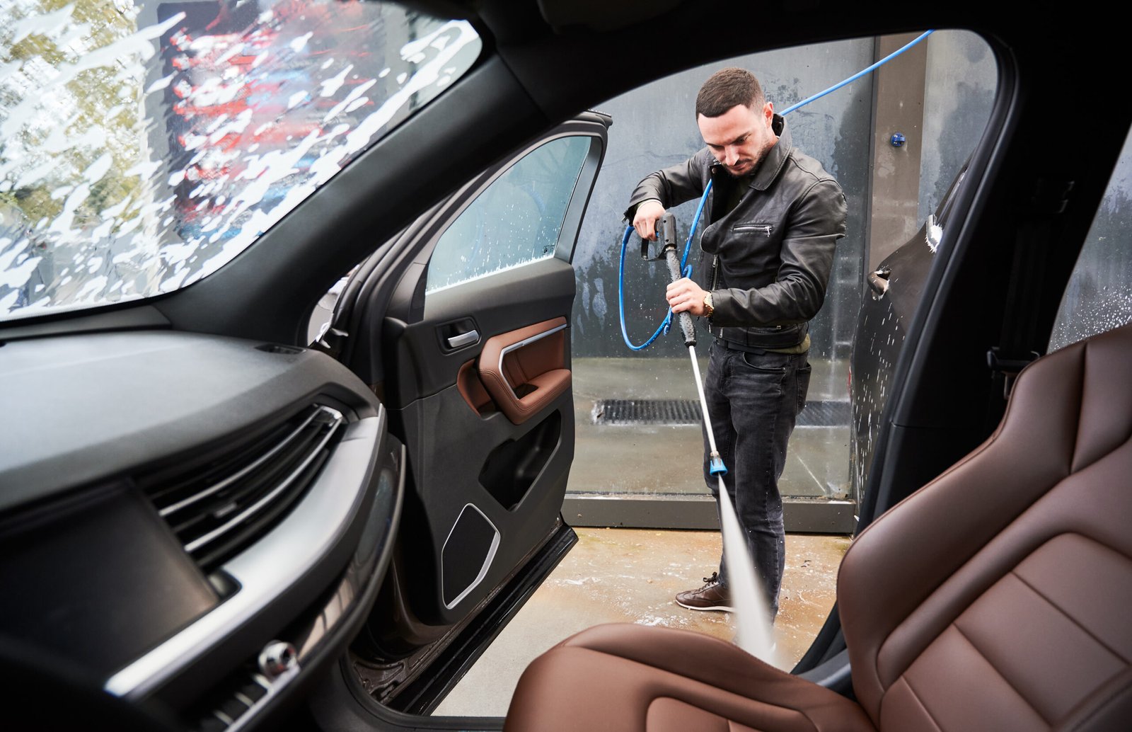 View from inside a car on young man washing car on carwash station outdoor. Handsome worker cleaning automobile, using high pressure water.
